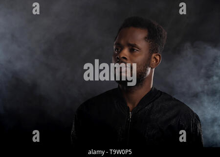 Cool strong handsome young african man with beard wearing black windbreaker jacket looking away with serious expression posing against white smoke on  Stock Photo