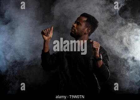 Waist up shot of handsome confident young african american man in black jacket with beard gesturing as dancing, raising hands up, looking to side seri Stock Photo