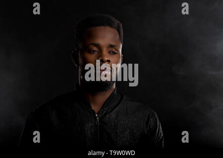 Portrait of attractive young afro man with small beard wearing black jacket looking away with sad and thoughtful expression on dark background with co Stock Photo