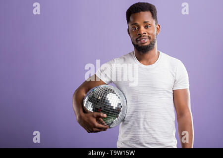 Happy dark skinned man holding shining disco ball, going to party with friends, celebrating something, wearing casual clothes, smiling standing over p Stock Photo
