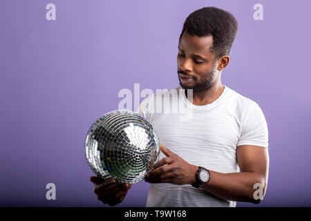 Happy charming dark skinned student holding shining silver disco ball, going to party with friends, celebrating pass exams wearing casual clothes, smi Stock Photo