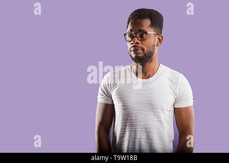 Portrait of intelligent calm young bearded black man in eyewear, dressed in casual clothes looking with puzzlement aside standing against purple studi Stock Photo