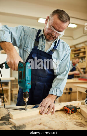 Portrait of mature carpenter drilling wood while working at table in joinery, copy space Stock Photo