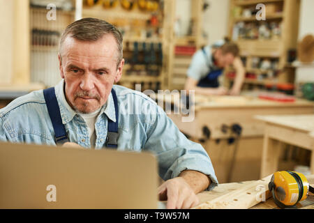 Portrait of senior carpenter using laptop while working in joinery, copy space Stock Photo