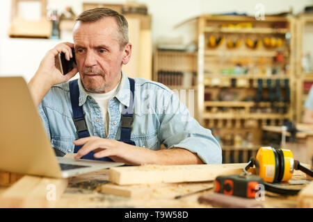 Portrait of modern senior carpenter speaking by phone and using laptop while working in joinery, copy space Stock Photo