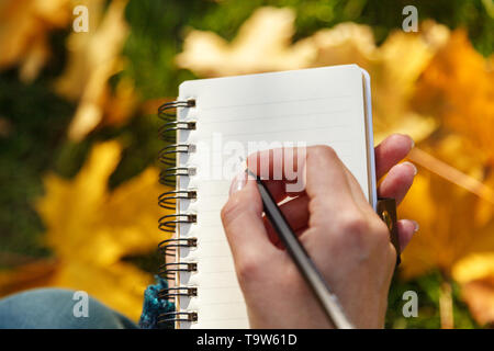 A woman's hand holding a paper notebook and pencil against the yellow leaves in the soft sunlight. Autumn mood, empty space for text Stock Photo