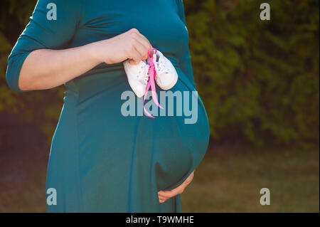 Small shoes for the unborn baby in the belly of pregnant woman. Pregnant woman holding small baby shoes relaxing at home in bedroom. Small shoes for Stock Photo