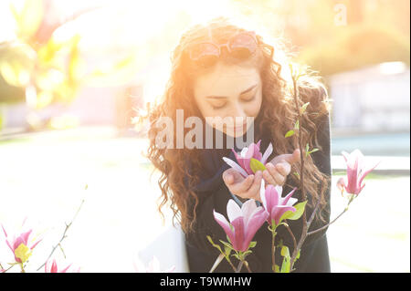 Young woman near blossoming magnolia flowers tree in spring park on sunny day. Beautiful happy girl enjoying smell in a flowering spring garden Stock Photo