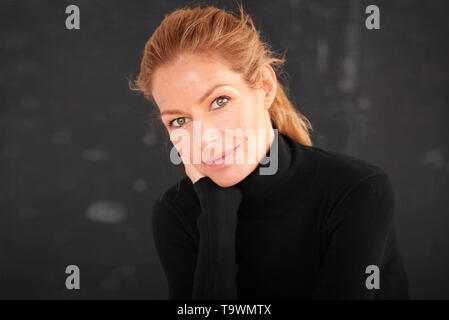 Portrait of beautiful mature woman wearing roll neck sweater and looking at camera while sitting with hand on chin at dark background. Stock Photo