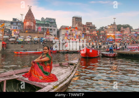 Young Indian female tourist in traditional dress (saree) on a wooden boat overlooking the historic Varanasi city architecture and Ganges river ghat at Stock Photo