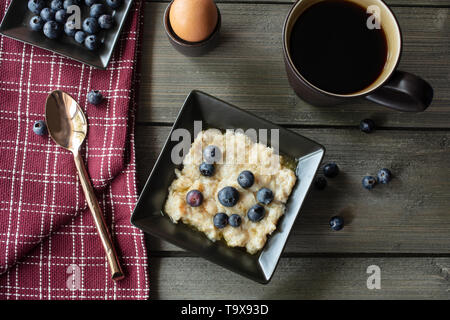 Breakfast bowl of oatmeal, honey, butter and blueberries with coffee and soft boiled eggs on a rustic table with a spoon and dish towel Stock Photo