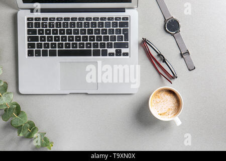 Laptop with cup of coffee, watch, eyeglasses and eucalyptus branch on grey table Stock Photo