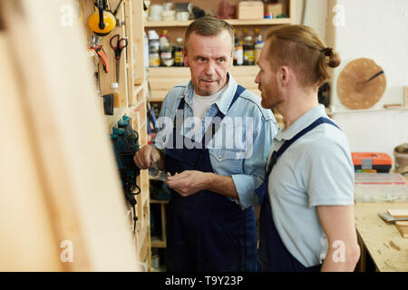 Portrait of senior carpenter talking to young trainee while working in joinery workshop, copy space Stock Photo