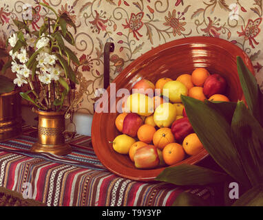 Oranges, apples and lemons in a clay bowl next to a vase with flowers. Stock Photo