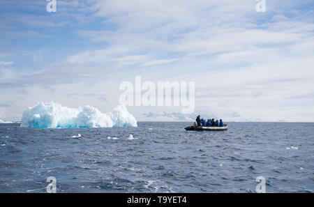 A group of tourists and a driver in a Zodiak boat cruising past a light and medium blue iceberg floating in the Southern Atlantic Ocean near Antarctic Stock Photo
