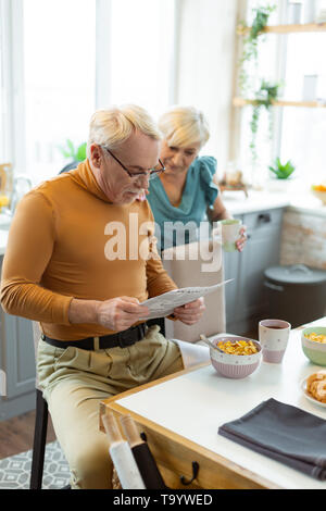 Focused nice-looking elderly white-haired being busy with a newspaper reading Stock Photo