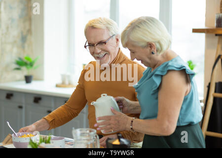 Kind lady pouring tea for her silver-haired attractive nice-looking spouse Stock Photo