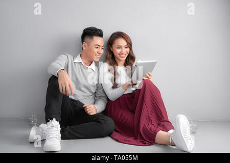 Happy young couple using tablet sitting on the floor at home isolated on gray background Stock Photo