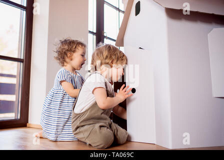 Two toddler children playing with a carton paper house indoors at home. Copy space. Stock Photo