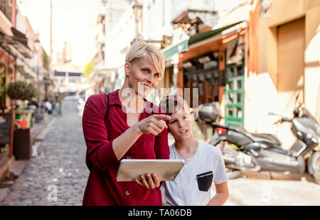 Mother with son standing outdoors in Mediterranean town, looking at map on tablet. Stock Photo