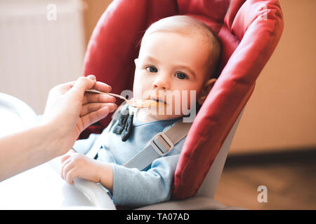 Baby boy eating mashed apple from spoon indoors. Looking at camera. Childhood. Stock Photo
