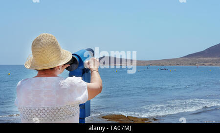 Rear view of senior woman traveler wearing hat and holding blue telescope binocular adjusting lens to see across the sea during summer holiday Stock Photo