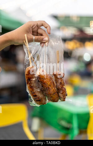 Sai Ua, the northen thai sausage in the hand of girl in a typical plastic bag on some wooden sticks, Thailand Stock Photo