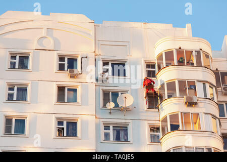 Industrial climber fixed on safety ropes puttying and painting facade of building on sunny day. Stock Photo