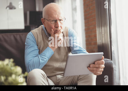 Senior man surprised using a digital tablet while sitting on couch at home Stock Photo
