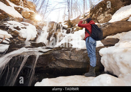 Beautiful girl in red jacket with black backpack stands arm up in front of the winter waterfall and orange sun Stock Photo