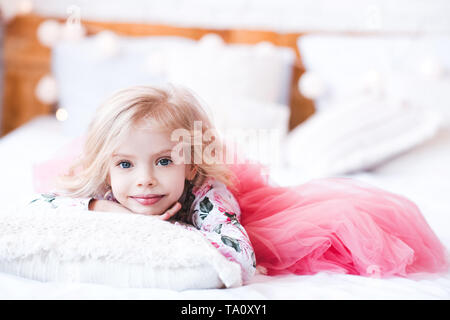 Little princess girl 3-4 year old wearing stylish fluffy dress lying in bed in room. Looking at camera. Childhood. Stock Photo