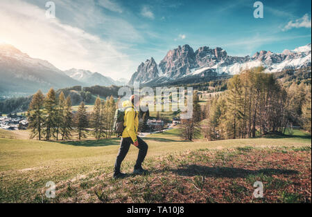 Young man in yellow jacket with backpack is walking on the green meadow against the mountains at sunset in spring. Landscape with sporty guy, snowy ro Stock Photo