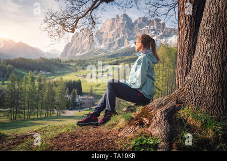 Beautiful young woman in sunglasses and blue jacket is sitting on the hill under the tree at sunset. Colorful spring landscape with sporty girl, meado Stock Photo