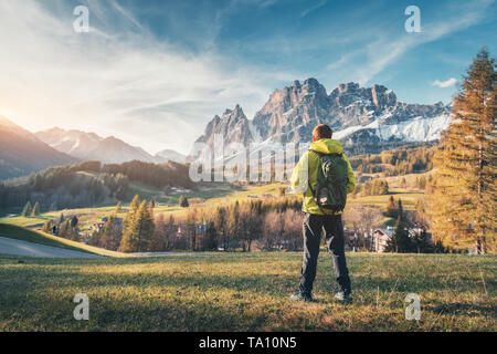 Young man in yellow jacket with backpack standing on the hill against the mountains at sunset in spring. Landscape with sporty guy, green meadow,  sno Stock Photo