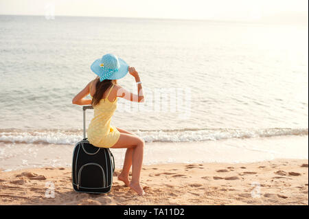 Travel holiday and people concept - woman tourist sitting near the sea on suitcase and in hat and watching sun and show thumbs up Stock Photo