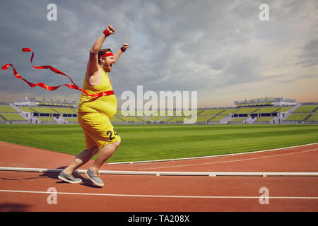 Fat funny man runs to the finish with a red ribbon on the race track in the stadium. Concept of winning motivation in health business. Weight loss. Stock Photo