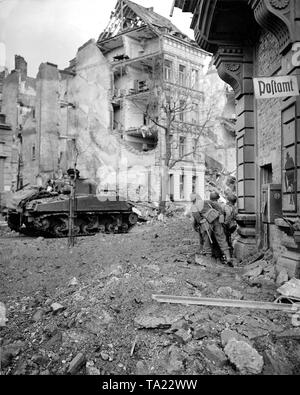 Infantrymen of the US Army (104th Division, 1st US Army) at a small distance from a tank (750th Battalion, 415th Infantry Regiment) peeking around a house corner. On the right, on a partially destroyed house facade a sign recalls a former post office. Stock Photo