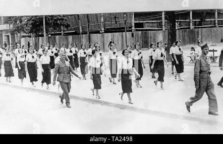 Photo of a group of marching women of Accion Ciudadana, a Spanish national civil movement, which was part of the CEDA (Spanish Confederation of Autonomous Right-wing Groups) until 1937. The photo was probably taken in Burgos, Castile and Leon, and shows women in white blouses and dark skirts with gorillo caps, on which a white cross was sewn. The women are accompanied by soldiers. On the left, an officer ranked comandante (Major). Stock Photo