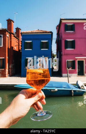 Hand of male holds glass with Spritz aperol cocktail overlooking colorful houses in Burano, Veneto, Italy