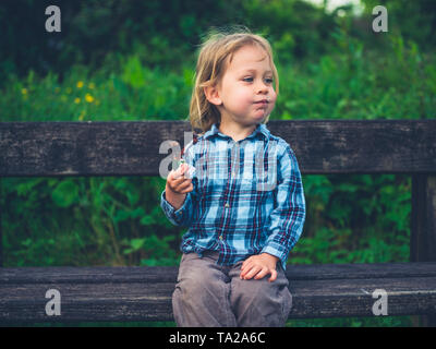 A little toddler is sitting on a bench in nature and is eating a fruit bar Stock Photo