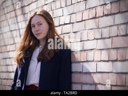 Red-haired beautiful girl in a blue coat stands near a brick wall with pink bricks, which illuminates the sunlight Stock Photo