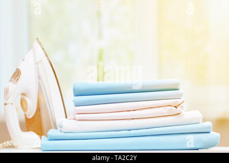 Close-up of blue and white clean bedding on a blurred background. A stack of folded bedsheets on the table, an iron stands nearby. Sunlight from the w Stock Photo