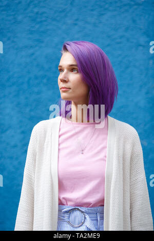 beautiful girl with violet hair on a blue background looks to the side and up, with an earring in her nose and a pendant around her neck, medium plan Stock Photo