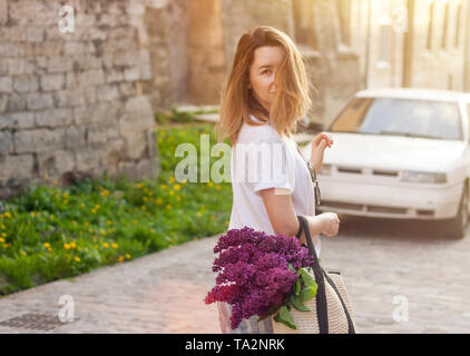 Stylish woman holding straw bag with a vivid bunch of lilac flowers and walking down the street. Moody sunny portrait. Stock Photo