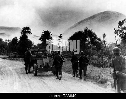 Soldiers march down a street in the Carpathians. A wagon pulls an infantry gun ( 7.5-cm-leichtes Infanteriegeschuetz 18) In the background, smoke clouds of artillery fire. Photo of the Propaganda Company (PK): War correspondent Voigt. Stock Photo