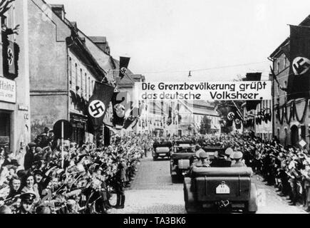 Part of Hitler's motorcade enters a place near the former German-Czechoslovak border on October 1, 1938. Above the street is a banner: 'The border population greets the German militia army.' On the left and right, cheering spectators. The car in the foreground belongs to the bodyguard of the SS. Stock Photo