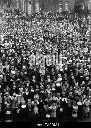In a schoolyard (presumably a girls' school) in Berlin, hundreds of schoolgirls have gathered with eating utensils in their hands to receive food donated by Quaker. Further behind are also some students and teachers. Stock Photo