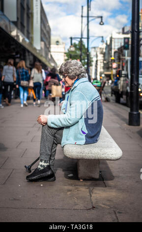 Side view portrait of an senior woman seated alone on a concrete bench, looking pensive, Oxford street, London, England, UK. Stock Photo
