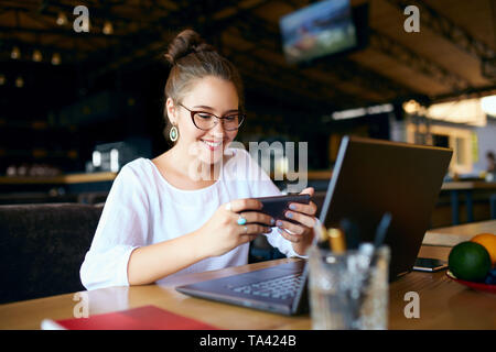 Mixed race business woman distracted from work on the laptop watching video on smartphone. Freelancer holding mobile phone and browsing using high spe Stock Photo