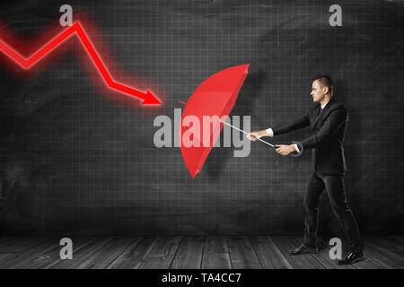 Businessman protecting himself with red umbrella against red diagram arrow on black background Stock Photo